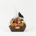 A gourmet snack and candy gift basket in a brown woven basket, wrapped in clear cellophane with a dark green bow.