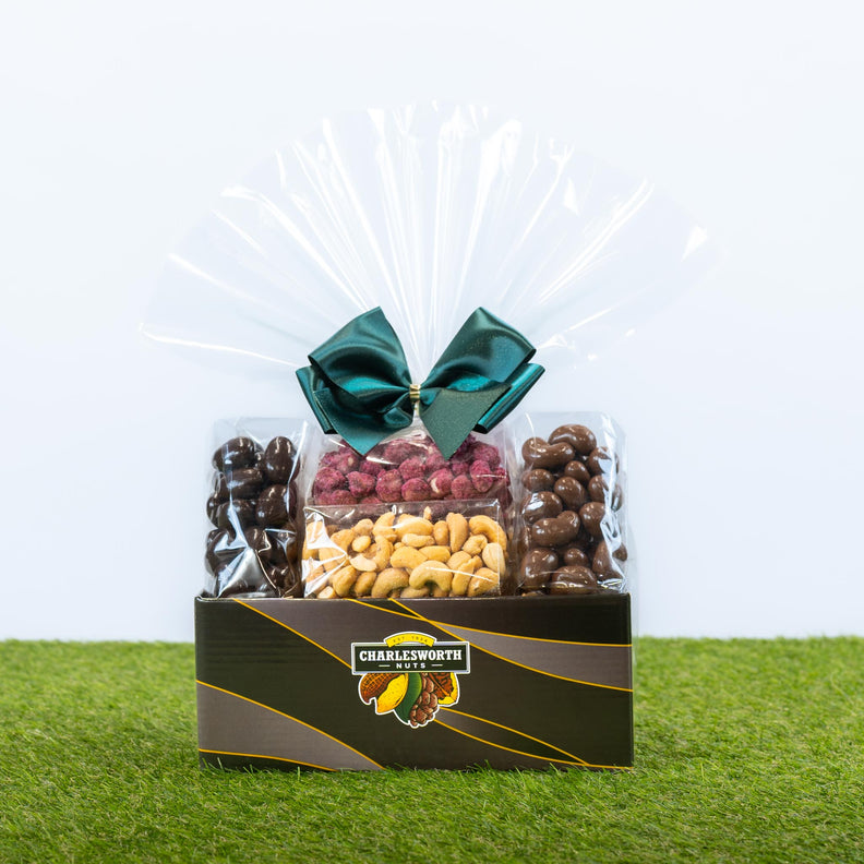 A gourmet snack gift hamper featuring chocolate-covered nuts and dried fruit in clear bags, wrapped in cellophane with a dark green bow, and presented in a branded black box.